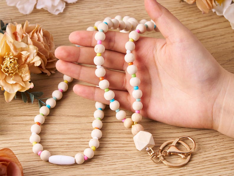 Pode incluir: Um cord&atilde;o de contas de madeira com um pingente geom&eacute;trico branco e ferragens douradas. O cord&atilde;o apresenta contas redondas de madeira de cor clara intercaladas com contas menores e coloridas em tons de rosa, laranja, amarelo, verde e azul.