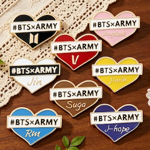 May include: A collection of heart-shaped enamel pins in various colors, including black, white, red, pink, yellow, brown, and blue. Each pin features the text "#BTSxARMY" and a name or initial. The pins are arranged on a decorative surface.