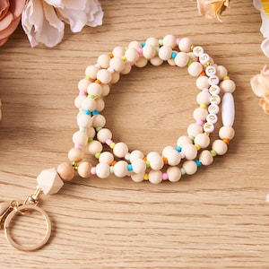 Pode incluir: Um cord&atilde;o de contas de madeira com fecho e chaveiro dourados. O cord&atilde;o apresenta contas de cor natural intercaladas com contas coloridas. Uma se&ccedil;&atilde;o do cord&atilde;o soletra "MS. CLAWSON" em contas brancas com letras douradas. O cord&atilde;o est&aacute; sobre uma superf&iacute;cie de madeira.