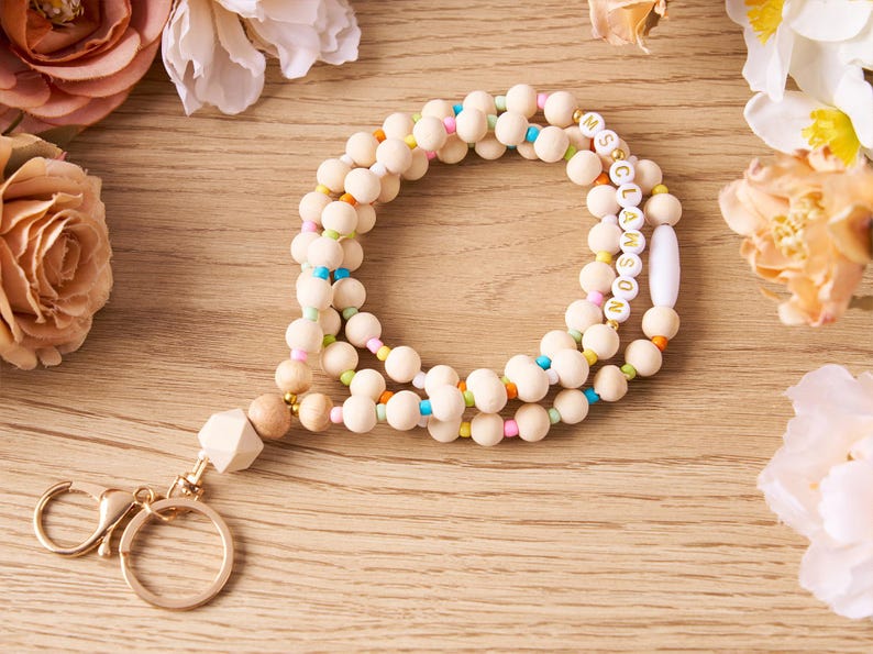 Pode incluir: Um cord&atilde;o de contas de madeira com fecho e chaveiro dourados. O cord&atilde;o apresenta contas de cor natural intercaladas com contas coloridas. Uma se&ccedil;&atilde;o do cord&atilde;o soletra "MS. CLAWSON" em contas brancas com letras douradas. O cord&atilde;o est&aacute; sobre uma superf&iacute;cie de madeira.