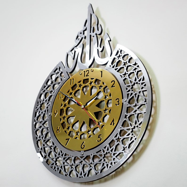 May include: A silver and gold wall clock with an intricate geometric design and the Arabic word "Allah" in the center. The clock face features a gold background with black Roman numerals and a starburst design.