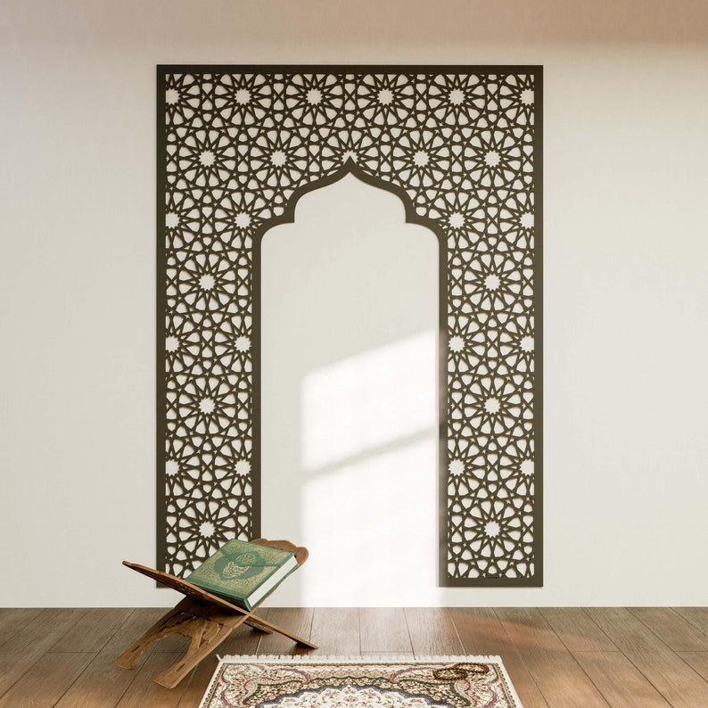 May include: A dark brown, intricately patterned wall panel with an arched top, resembling a traditional Islamic design. A wooden book stand holds a green book, and a patterned rug is on the wooden floor.