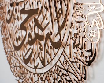 Op de afbeelding: Een close-up van een gouden metalen wandkunstwerk met ingewikkelde Arabische kalligrafie. Het ontwerp is rond en heeft een herhalend patroon van letters en symbolen.