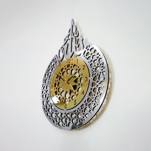 May include: A silver and gold wall clock with an intricate geometric design and the Arabic word "Allah" in a decorative font. The clock face features a gold background with black Roman numerals and a starburst design.