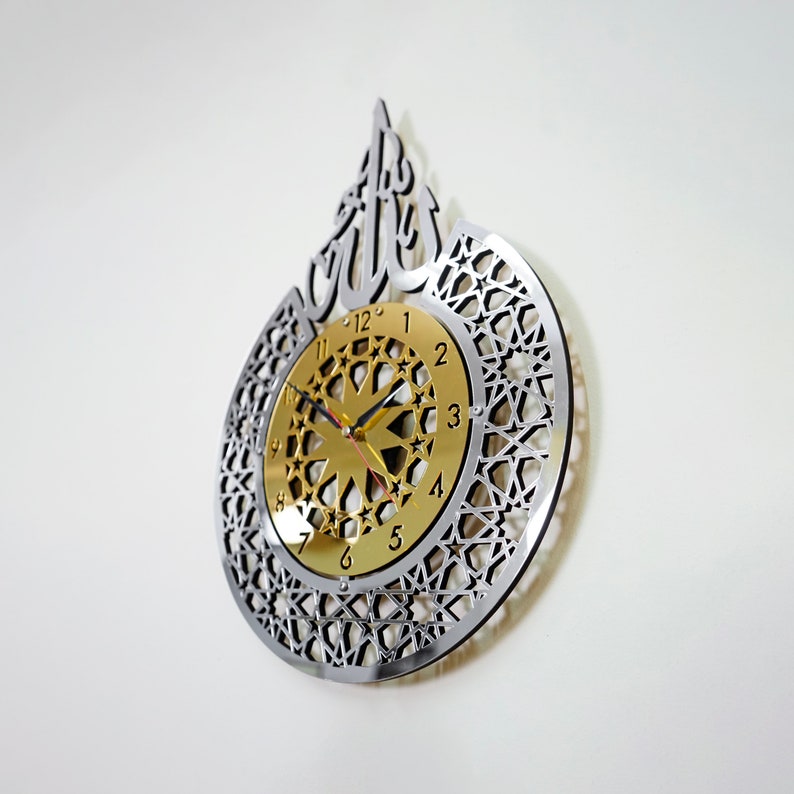 May include: A silver and gold wall clock with an intricate geometric design and the Arabic word "Allah" in a decorative font. The clock face features a gold background with black Roman numerals and a starburst design.