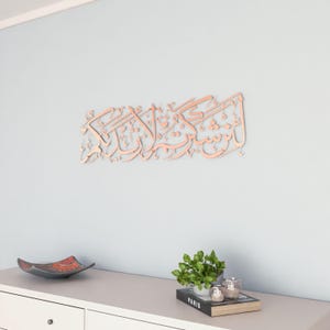 May include: A rose gold metal wall decor with Arabic calligraphy. The decor is hanging on a light blue wall above a white dresser with a book and a plant on top.