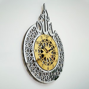 May include: A silver and gold wall clock with an intricate geometric design and Arabic calligraphy. The clock face features a gold background with black Roman numerals and a starburst pattern.