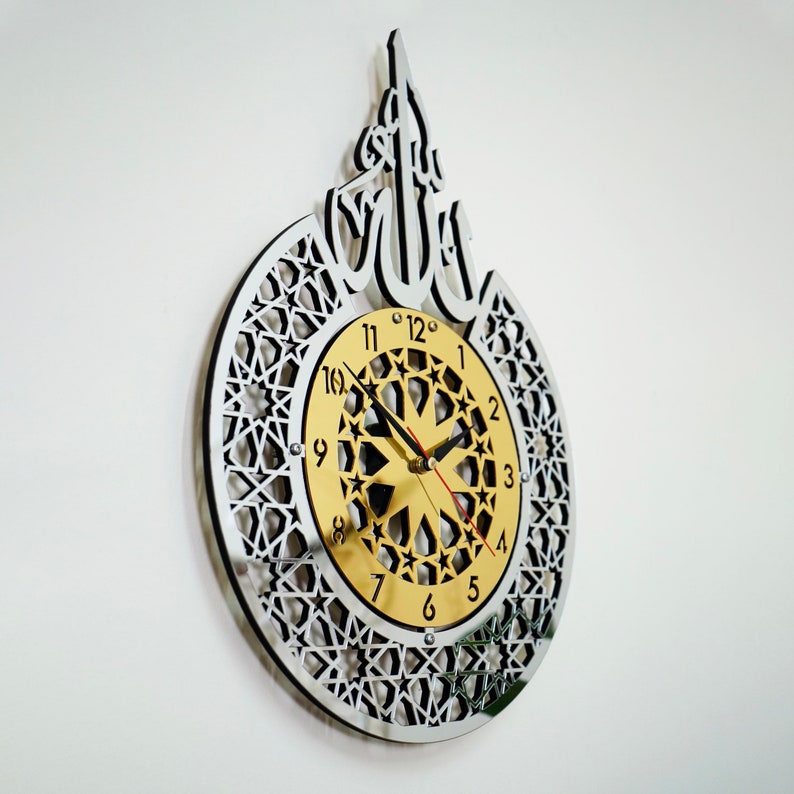 May include: A silver and gold wall clock with an intricate geometric design and Arabic calligraphy. The clock face features a gold background with black Roman numerals and a starburst pattern.