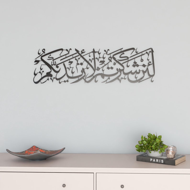 May include: Black metal wall art featuring Arabic calligraphy. The art is mounted on a white wall above a white dresser with two drawers. A small potted plant and a book titled "Paris" are on the dresser.