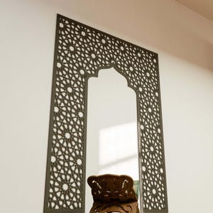 May include: A decorative wall mirror with a dark brown geometric pattern frame. The frame has an arched top and intricate star and floral designs. The mirror is rectangular with a clear reflection.