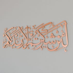 May include: A copper-colored metal wall art piece featuring Arabic calligraphy. The calligraphy is in a traditional style and is arranged in a flowing, decorative pattern.