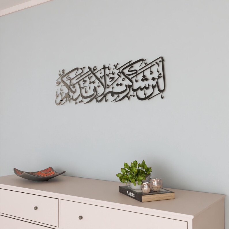 May include: A black metal wall art piece with Arabic calligraphy. The art is hanging on a light blue wall above a white dresser with two drawers. A small potted plant and two candle holders are on top of the dresser.