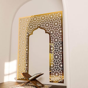 May include: A gold-coloured, geometric wall panel with an arched opening, featuring intricate star and floral patterns. A wooden book stand with an open book rests on a patterned rug in front of the panel. The wall is white.