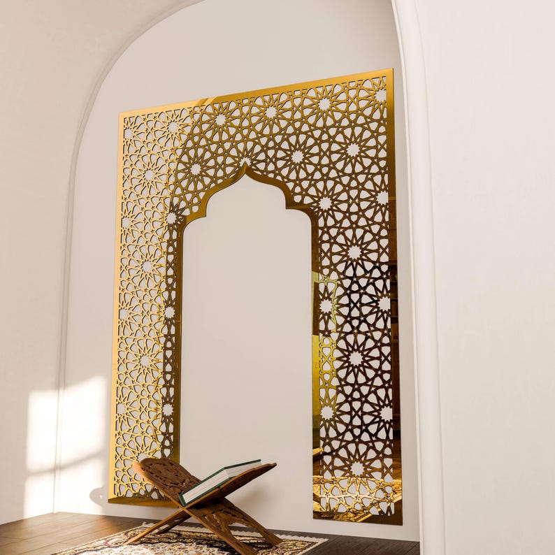 May include: A gold-coloured, geometric wall panel with an arched opening, featuring intricate star and floral patterns. A wooden book stand with an open book rests on a patterned rug in front of the panel. The wall is white.