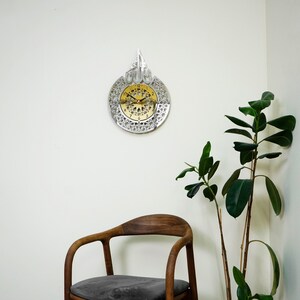 May include: A silver and gold clock with an intricate design. The clock is hanging on a white wall with a wooden chair and a green plant in the background.