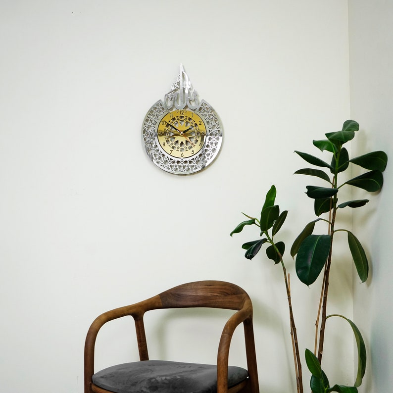 May include: A silver and gold clock with an intricate design. The clock is hanging on a white wall with a wooden chair and a green plant in the background.
