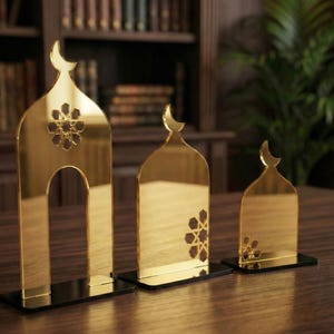 Op de afbeelding: Drie goudkleurige decoratieve stukken in de vorm van boogvormige deuropeningen met een maan erbovenop. Elk stuk heeft een geometrisch bloemmotief en staat op een zwarte basis. Ze staan op een houten oppervlak.