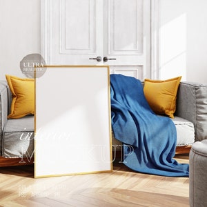 May include: A gold framed picture mockup in a living room setting. The picture is leaning against a grey sofa with a blue throw blanket and yellow cushions. The floor is wooden.
