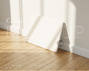 Könnte beinhalten: Ein weißer Leinwand-Mockup, der an einer weißen Wand mit Holzboden lehnt. Die Leinwand ist leer und bereit für Ihr Design. Der Text "Canvas Mockup" ist in Weiß an die Wand geschrieben.