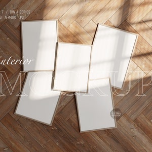 May include: Five blank picture frames in various sizes are arranged on a wooden floor. The frames are all white with a thin, light brown border. The floor is made of light brown wood planks arranged in a herringbone pattern. The frames are arranged in a staggered pattern, with the largest frame in the center.