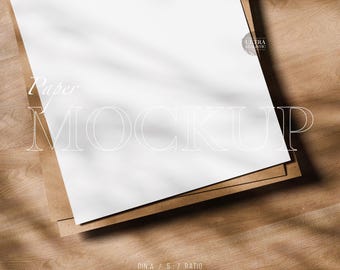 May include: A white paper mockup with the word "Paper" and "MOCKUP" in a stylish font. The paper rests on a brown cardboard base, set against a wooden background. The image includes the text "DIN A / 5:7 RATIO PSD AFPHOTO JPG".