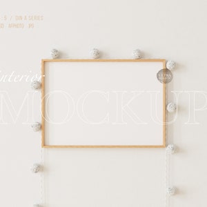 Puede incluir: Una maqueta de marco de madera con fondo blanco, colgada en una pared blanca. Una cadena de luces blancas con pequeños pompones blancos está drapeada alrededor del marco. El texto "interior mockup" está escrito en blanco en la pared.