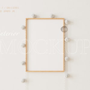 Puede incluir: Una pared blanca con una maqueta de marco de madera, que presenta un fondo blanco y una cadena de pompones blancos colgando de la pared. El texto "interior mockup" está escrito en blanco en la pared.