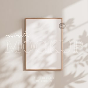 Puede incluir: Una maqueta minimalista con un marco de madera marrón claro en blanco sobre un fondo blanco. El marco está colocado contra una pared blanca con luz natural suave y sombras proyectadas por una ventana.