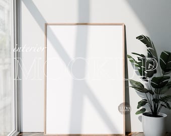 May include: A large, blank white picture frame with a light wood border leans against a white wall. A potted plant with green leaves sits to the right. The words "interior MOCKUP" are printed on the frame.