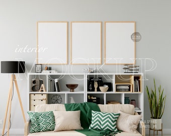 Op de afbeelding: Een woonkamer interieur mockup met drie lege frames aan de muur boven een wit boekenrek met decoratieve items. Een beige bank met groene kussens en een groene deken bevindt zich op de voorgrond.