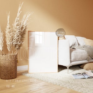 May include: A white sofa with pillows and a throw blanket sits in a room with a beige wall and a wooden floor. A large, framed picture sits on the floor in front of the sofa. A vase with dried pampas grass sits on the floor to the left of the sofa.