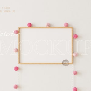 Puede incluir: Un marco de madera con fondo blanco, colgado en una pared blanca. Una cadena de pompones rosas y blancos cuelga alrededor del marco. El texto "interior mockup" está escrito en blanco en la pared.