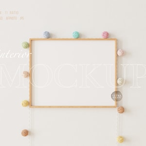 Puede incluir: Un marco de madera con fondo blanco cuelga de una pared blanca. Una cadena de luces de bolas de algodón de color pastel cuelga por encima y a la derecha del marco. El texto "interior mockup" está escrito en blanco en la pared.