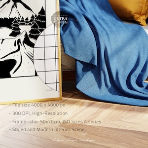 May include: A framed black and white illustration of a mountain scene with trees. The frame is gold and the illustration is on a white background. The frame is sitting on a wooden floor with a blue blanket draped over a white sofa.