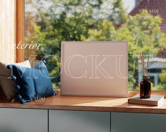 May include: A white picture frame mockup on a wooden shelf with a blue pillow, a book, and a diffuser with reeds. The frame is sitting in front of a window with rain drops on the glass.