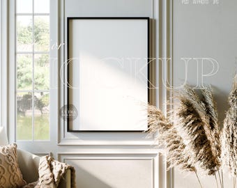 May include: A black framed vertical poster mockup with a white interior. The mockup is in a room with a window, a beige couch, and decorative pampas grass. The text "Interior Mockup" is overlaid on the image.