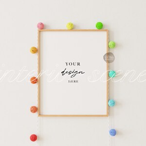 K&ouml;nnte beinhalten: Ein wei&szlig;er Rahmen mit Holzrahmen h&auml;ngt an einer wei&szlig;en Wand. Der Rahmen ist von einer Schnur mit bunten Baumwollkugeln umgeben. Der Text "YOUR design HERE" ist in Schwarz im Rahmen geschrieben.