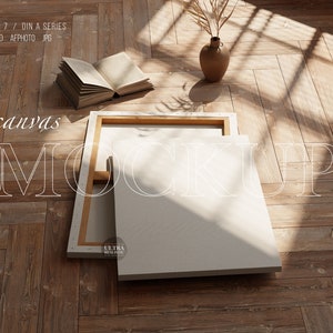 May include: A white canvas mockup with a wooden frame, lying on a wooden floor with a book and a vase with dried flowers. The text "canvas mockup" is written on the floor in white letters. The text "ultra realistic" is written on the canvas.