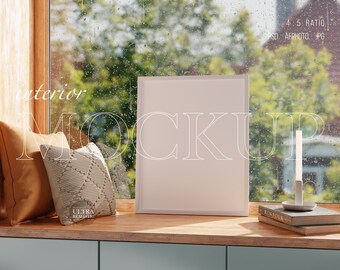 May include: A white picture frame mockup on a wooden shelf with two pillows, a candle, and books. The frame is sitting in front of a window with a blurry view of trees and rain.