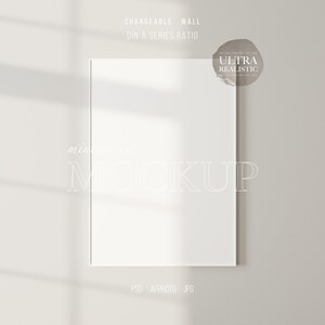 May include: A white frame mockup on a light grey wall with a shadow from a window. The text "CHANGEABLE WALL DIN A SERIES RATIO" is above the frame and the text "ULTRA REALISTIC" is in a circle to the right of the frame. The text "minimalist MOCKUP" is below the frame.