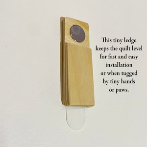 May include: A wooden ledge with a magnetic backing, designed to keep a quilt level during installation. The ledge is attached to a white wall with a sticky adhesive.
