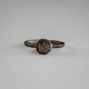 May include: A dark, weathered metal ring featuring a central octagonal face. The face is engraved with a distinct monogram consisting of the letters K and H intertwined. The band of the ring shows a textured, aged patina with visible signs of wear.