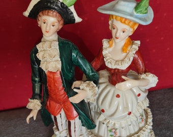 May include: A porcelain figurine set of a couple in 18th-century attire. The man is wearing a green jacket, red pants, and a white hat with a green feather. The woman is wearing a red dress with a white lace trim and a blue hat with a green feather. The figurines are standing on a white base with green and gold accents.