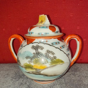 May include: A white porcelain sugar bowl with a red and blue lid. The bowl has a hand-painted scene of a mountain range with trees and a river. The bowl has two handles and a chipped lid.