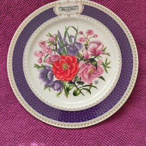 Puede incluir: Un plato de cerámica blanco con un borde dorado y morado. El centro del plato presenta un diseño floral con flores rosas, rojas, moradas y verdes. El plato tiene una pequeña imagen de un edificio en el centro superior.