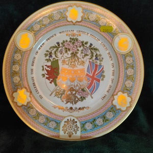 Puede incluir: Un plato conmemorativo de cerámica blanca con un borde dorado y un borde floral. En el centro se encuentra un escudo de armas con un dragón, un león y una bandera de la Union Jack. El texto dice "To Celebrate the Birth of HRH Prince William Arthur Philip Louis Born on the 21st June 1982".
