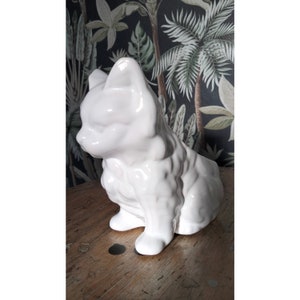Op de afbeelding: Een glanzend witte keramische kattenfiguur. De kat zit met zijn hoofd opzij, waarbij zijn oren en gezichtskenmerken te zien zijn. Het beeldje heeft een getextureerd lichaam en staat op een houten oppervlak.
