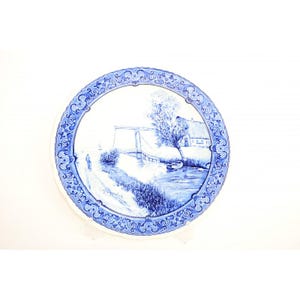 Op de afbeelding: Een ronde, witte keramische plaat met een blauw-witte landschapsafbeelding. De afbeelding toont een brug over een rivier, een huis en bomen. De plaat heeft een decoratieve blauwe rand.