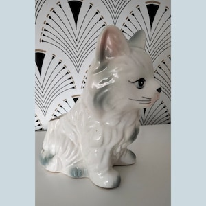 Pode incluir: Vaso de cerâmica branco em forma de gato com um design de pelo cinza e branco. O gato está sentado com a cabeça virada para o lado e tem um nariz rosa e olhos pretos.