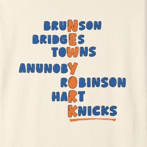 May include: A cream-colored short-sleeve t-shirt featuring blue text listing the names Brunson, Bridges, Towns, Anunoby, Robinson, Hart, and Knicks, with the word NEW YORK spelled vertically in orange letters down the center of the design.